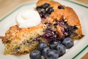A slice of blueberry coffee cake topped with a dollop of whipped cream sits on a plate next to several fresh blueberries.
