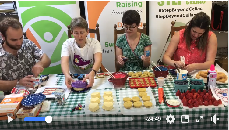 Four people sit at a table covered with a green checked cloth, decorating cupcakes and heart-shaped cakes. Baking supplies, fruit, and bowls of icing are on the table. Banners about celiac awareness are in the background.