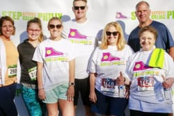 A group of seven people pose together outdoors in front of a “Step Beyond Celiac Philly” event backdrop. Some wear event t-shirts and bib numbers, with one woman giving a thumbs up.