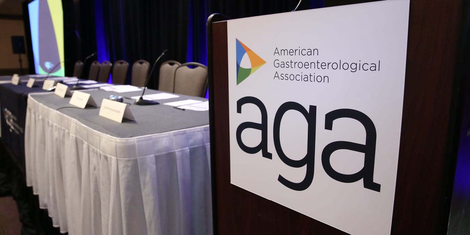 A conference table with several chairs, microphones, and name placards is set up on a stage. A sign in the foreground displays the American Gastroenterological Association (AGA) logo.