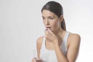 A woman with dark hair in a ponytail holds a glass of water in one hand and is about to take a pill with the other. She wears a light gray tank top and looks ahead with a neutral expression.