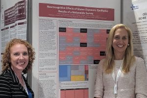 Two women stand in front of a scientific poster titled “Neurocognitive Effects of Gluten Exposure: Qualitative Results of a Nationwide Survey.” The poster contains text and a multicolored data chart.