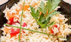 A close-up of a rice salad with chopped red and orange bell peppers, garnished with fresh parsley and chives, served in a black bowl.