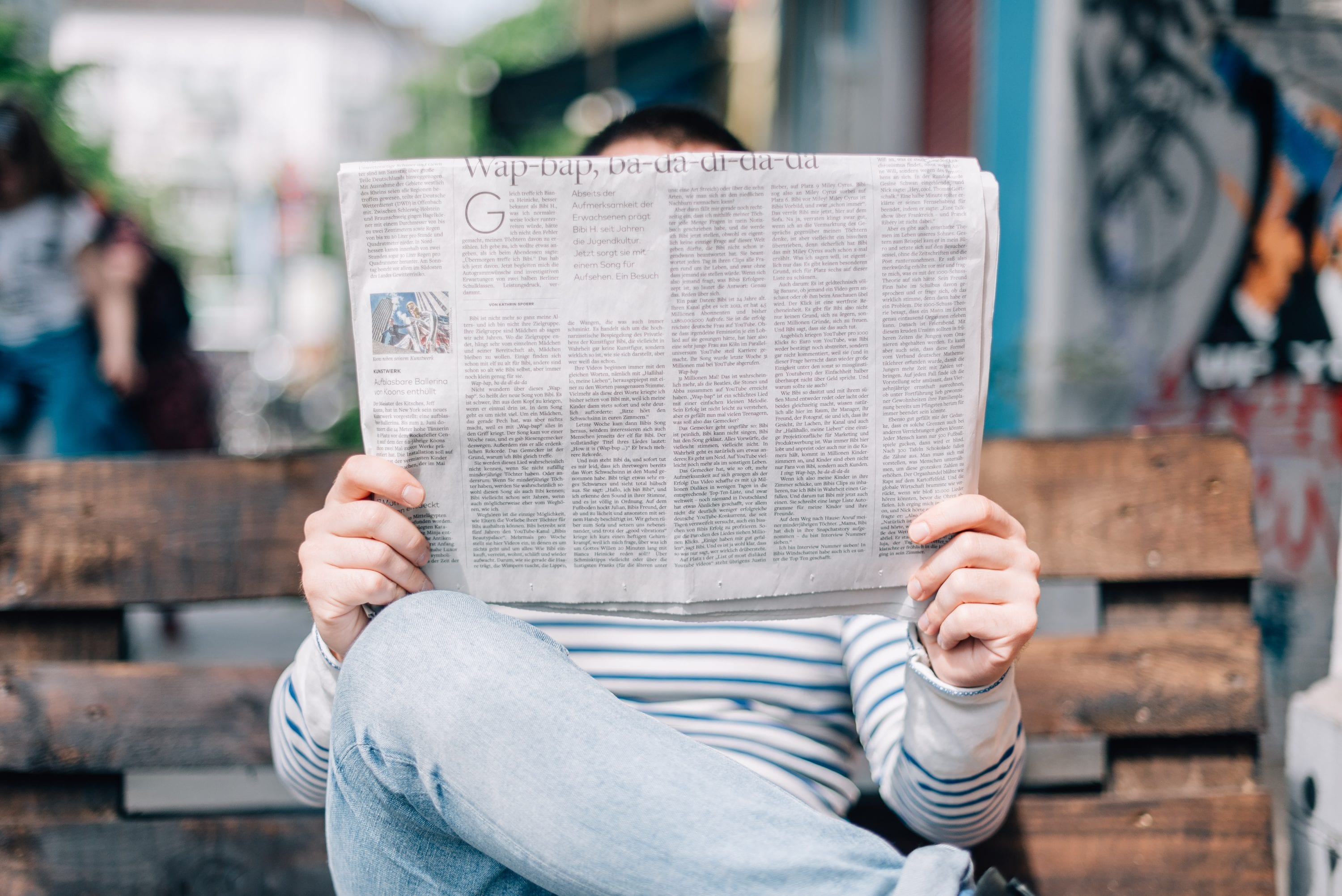 A person sitting on a wooden bench holds an open newspaper, obscuring their face. The background is slightly blurred, showing an outdoor urban setting with people and graffiti.