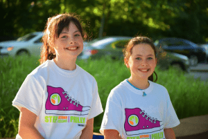 Two girls with light skin and brown hair wearing white Step Beyond Celiac Philly t-shirts stand outdoors on a sunny day, with cars and green foliage in the background. They are both smiling at the camera.
