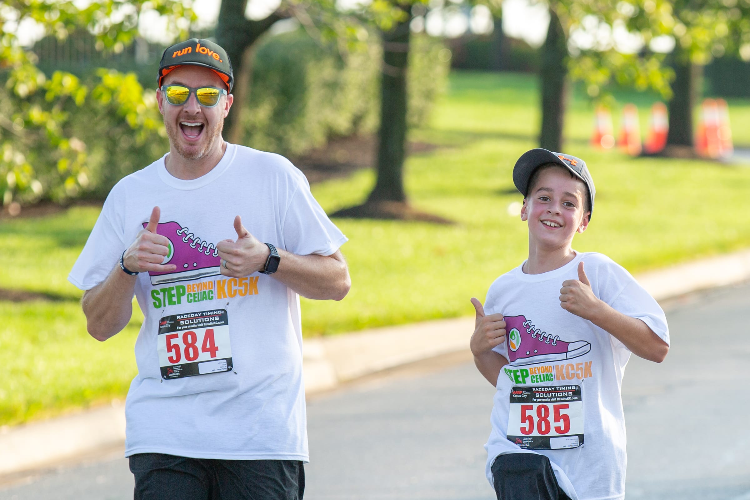 An adult and a child wearing matching white race t-shirts and numbered bibs give thumbs up while running outdoors on a sunny day. Trees and orange cones are visible in the background.