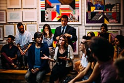 A group of people sit and stand in an art gallery, having a discussion. Colorful abstract paintings hang on the walls behind them. One woman in the center speaks while others listen.