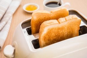Two slices of toasted bread are in a white toaster on a wooden surface. In the background, there is a cup of black coffee, a small dish of yellow jam, and a folded napkin.