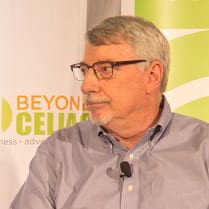 A man with gray hair, glasses, and a trimmed beard wears a light blue button-down shirt and sits in front of a Beyond Celiac banner, looking to his left. A microphone is clipped to his shirt.