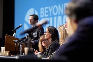 A panel of people speaks at a conference table with microphones, while a person stands at a podium in the background. The screen behind them displays the words Beyond and part of another word.