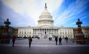 The United States Capitol building with its white dome, surrounded by a few people walking and taking photos in the foreground on a clear day.
