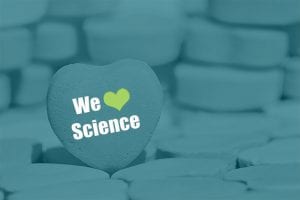 A heart-shaped tablet in the foreground displays the words “We [heart] Science,” with a heart symbol replacing the word “love.” Blurred, round tablets are stacked in the background. The image has a blue-green tint.