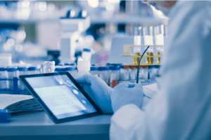 A person in a lab coat and gloves uses a tablet in a laboratory setting, surrounded by test tubes and laboratory equipment, with scientific data displayed on the tablet screen.