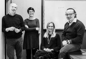 Four adults pose in a classroom. Two people stand while two are seated. There is a whiteboard with faint writing in the background. All are dressed in casual or business casual attire and look toward the camera.