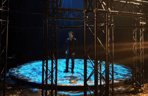 A person stands on a circular, illuminated platform surrounded by black metal scaffolding, with blue and yellow light patterns projected on the floor. The background is dark and the scene appears to be indoors.