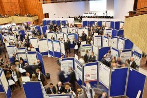 Large indoor scientific conference with many people walking among blue display boards showing posters. Attendees are reading, discussing, and presenting research in a spacious hall with a stage at the front.