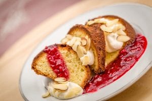 Two slices of bundt cake with whipped cream, sliced almonds, and a red fruit sauce are served on a white plate.