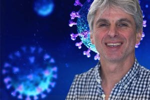 A man with gray hair and a checkered shirt smiles at the camera, with a digitally rendered background showing enlarged images of viruses colored blue and purple.