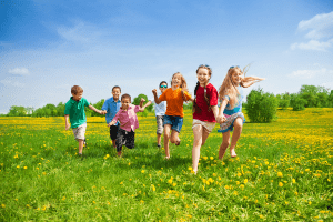 A group of seven children run hand in hand through a green field with yellow flowers under a blue sky. The children are smiling and dressed in colorful summer clothes. Trees and grass extend into the background.