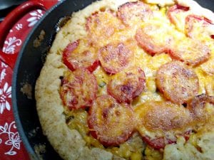 A close-up of a baked pie in a pan with a golden, flaky crust, topped with sliced tomatoes and melted cheese. The pie sits on a red and white floral-patterned cloth.