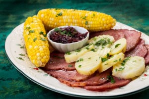 Sliced ham on a plate with potatoes and corn on the cob as sides