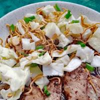 A plate of grilled meat slices topped with shredded cabbage, crispy noodles, and chopped green onions on a light-colored dish.