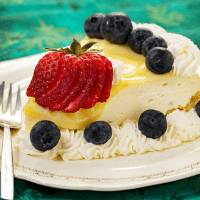 A slice of cheesecake topped with a sliced strawberry and several blueberries, with whipped cream and additional blueberries around the edge, on a white plate with a fork.