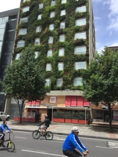 A building covered in vines and plants. People are biking on the street in front of the building. 