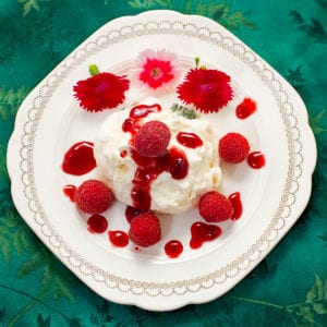Snow ball on a plate with raspberries and red flowers surrounding it.
