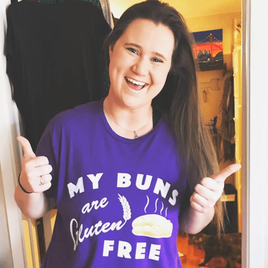 A photo of Katy smiling with her thumbs up. Her shirt says "My buns are gluten free"