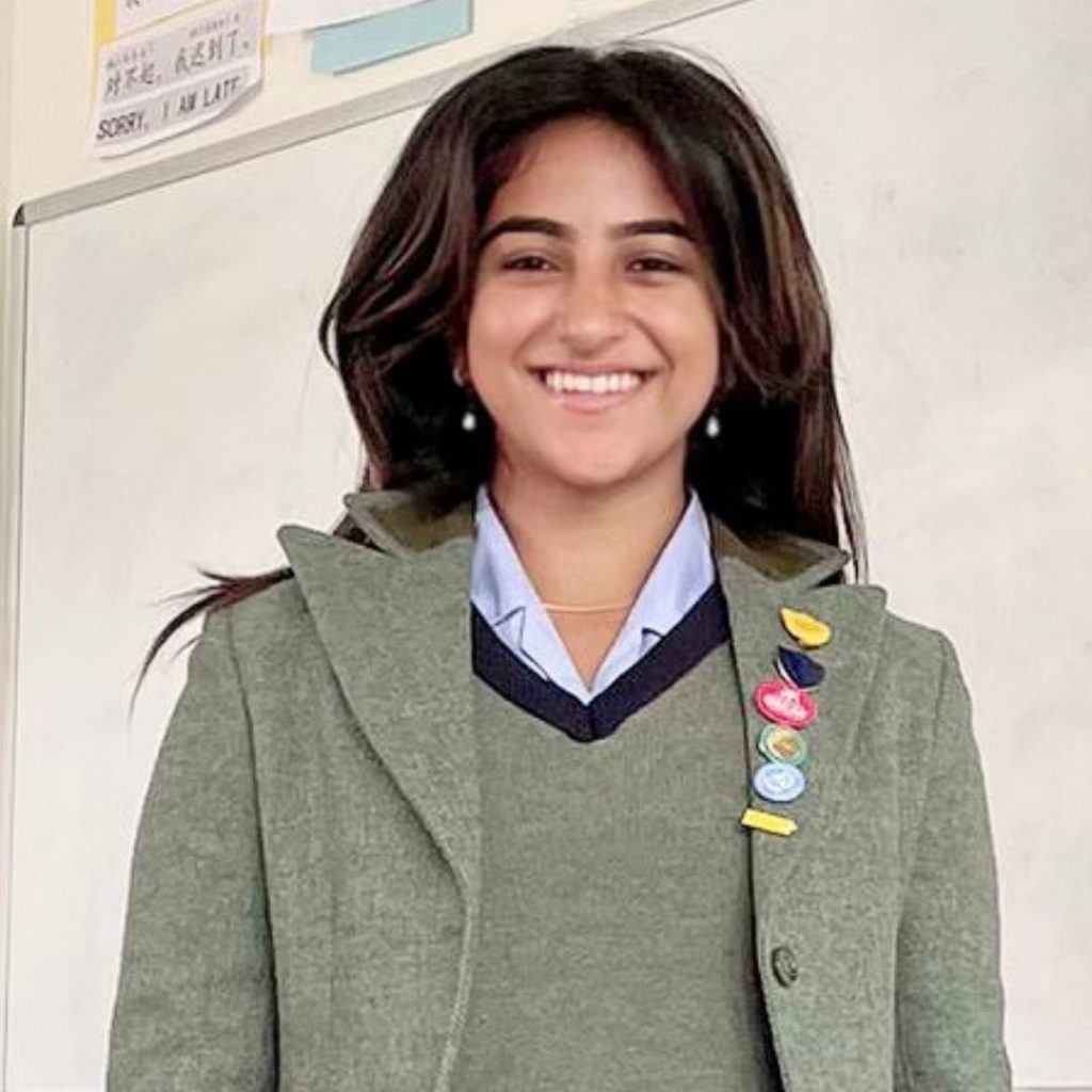 A person with long dark hair smiles while standing in front of a whiteboard, wearing a green blazer and sweater with several colorful pins on the lapel, and a collared shirt underneath.