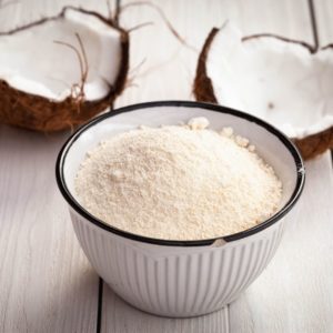 A bowl of coconut flour in front of two halves of a coconut. 