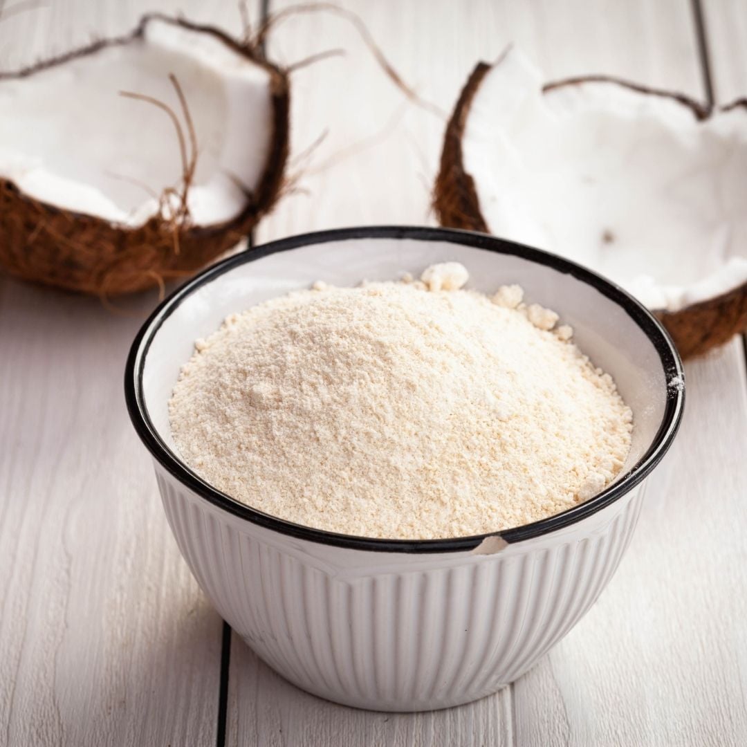 A bowl of coconut flour in front of two halves of a coconut.