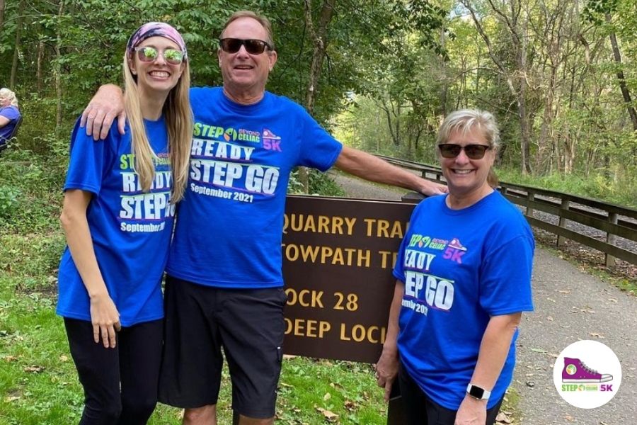 A photo of three people wearing the September 2021 Step Beyond Celiac 5K shirt. They are posing in front of a woodland trail. 