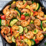 A photo of a skillet filled with shrimp, sausage, zucchini and peppers.