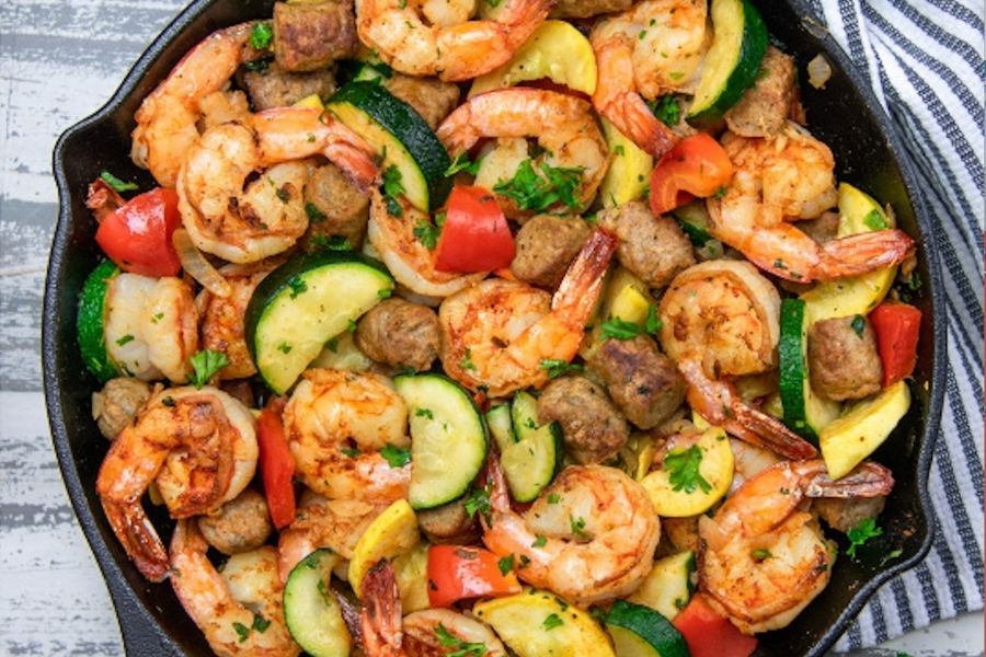 A photo of a skillet filled with shrimp, sausage, zucchini and peppers.