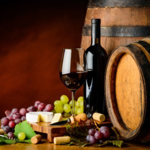 A glass of wine next to grapes, cheeses, and casks of wine.