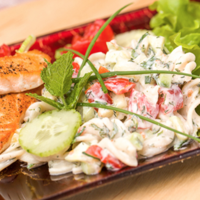 A plate with grilled salmon, a creamy pasta salad with tomatoes, herbs, and cucumbers, garnished with a mint sprig and chives, and a side of leafy green lettuce.