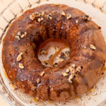 The apple cake on top of a plate. It was baked in a bundt pan. Caramel and walnuts top it. 