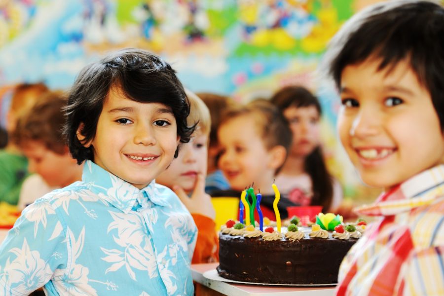 Children smiling in a classroom. Two sit in front of a chocolate cake. 