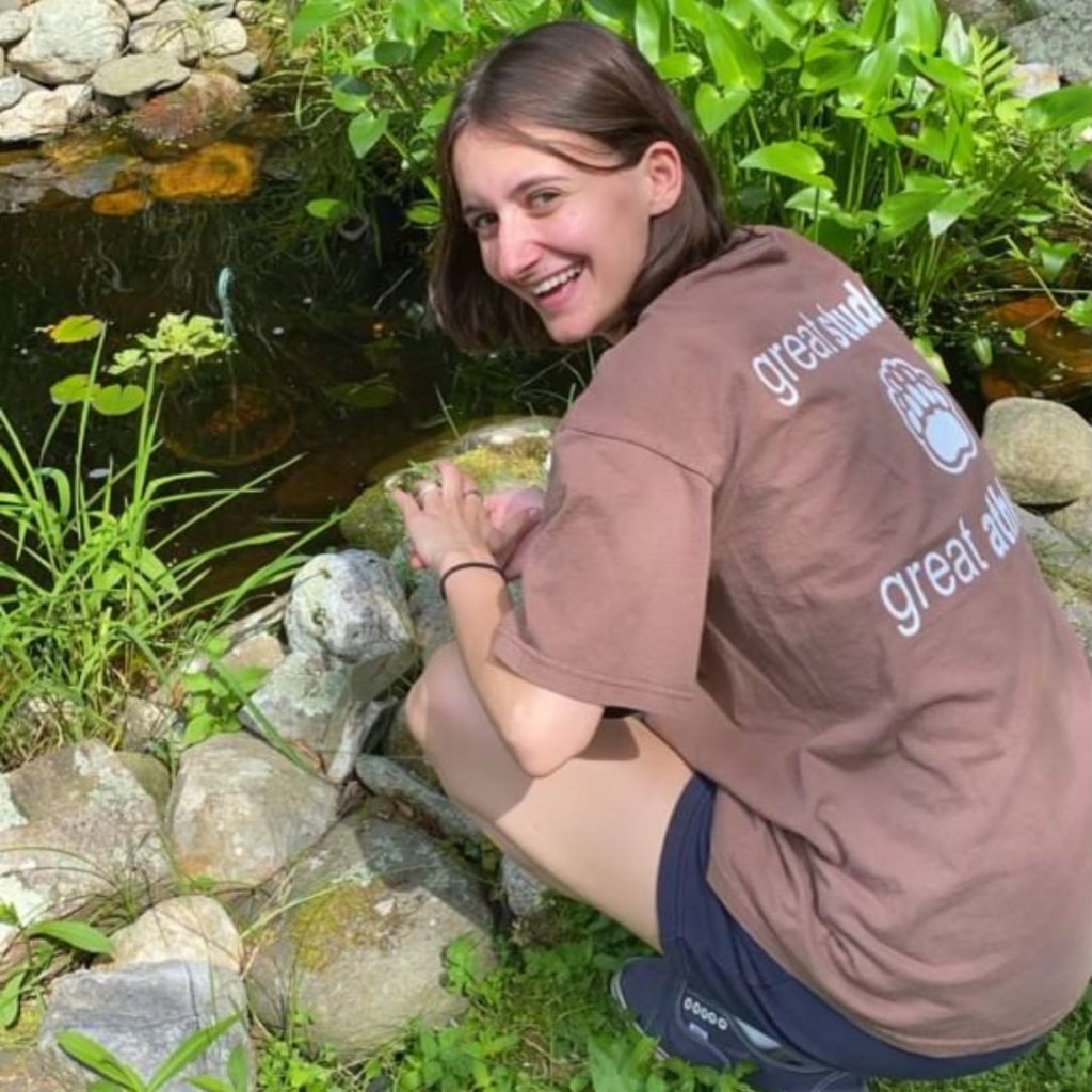 A photo of Leah kneeling by a pond, looking back at the camera and smiling.