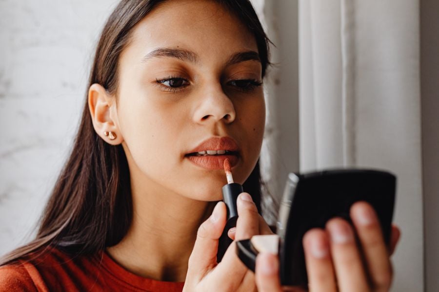 A person with long brown hair applies lipstick while looking into a compact mirror. They are wearing a red shirt and have small earrings in both ears.