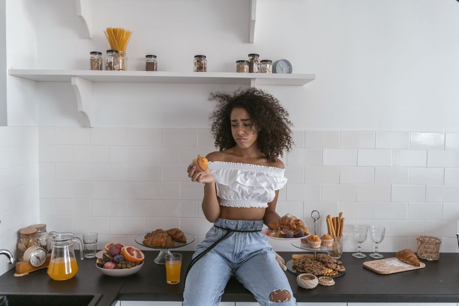 A woman sitting on a counter surrounded by pastries and breads. She is frowning sadly at a muffin in her hand. 