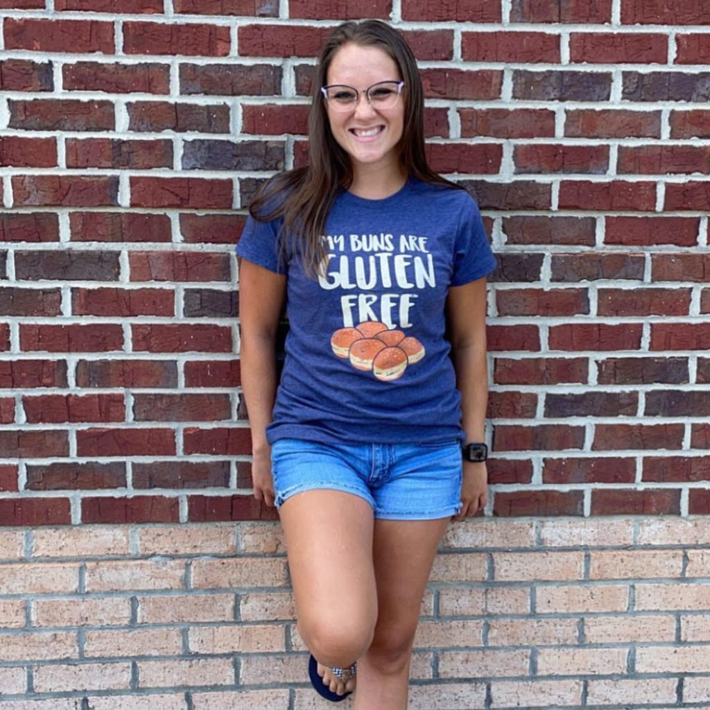 A photo of Elana. She is smiling and leaning against a brick wall. Her shirt says, "These buns are gluten-free."