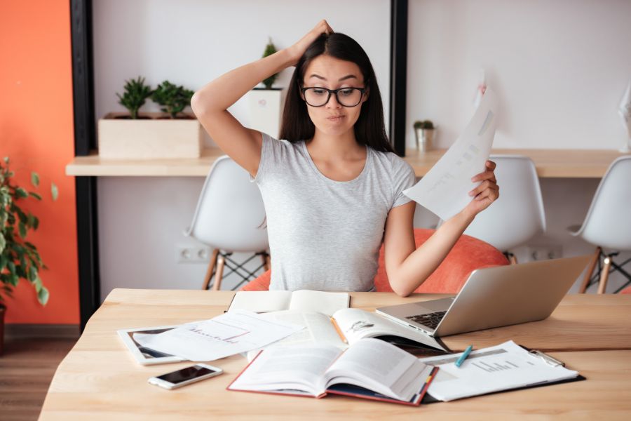 A photo of a woman sitting in front of a laptop and lots of paper, looking confused.