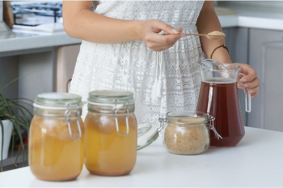 A person putting a spoon into a pitcher of some juice. Other jars are on on the counter.