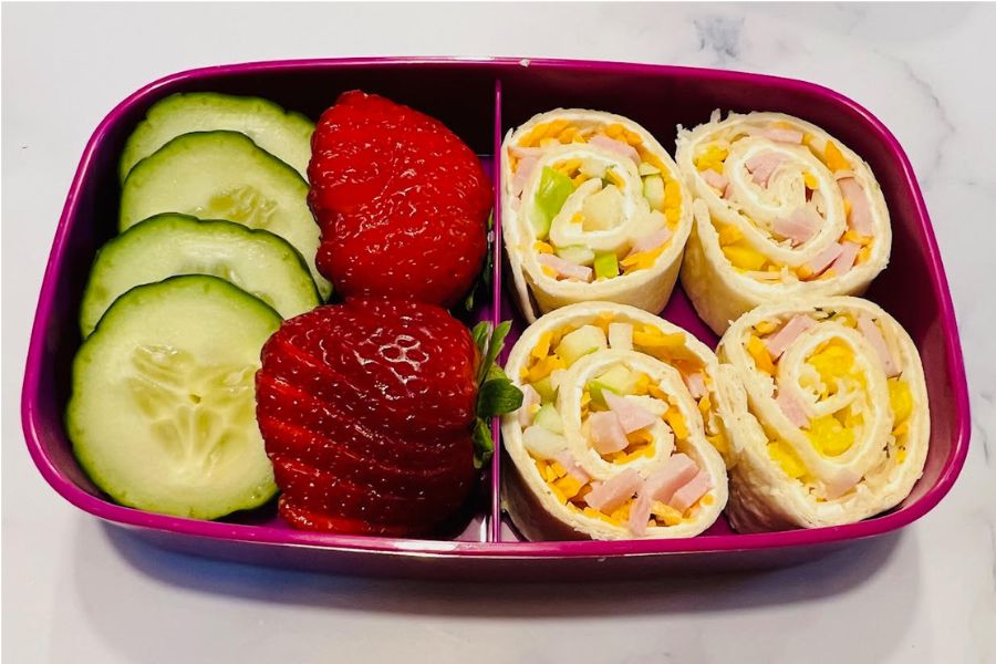 A lunchbox with the pinwheels on one side and cucumbers and strawberries on the other. 