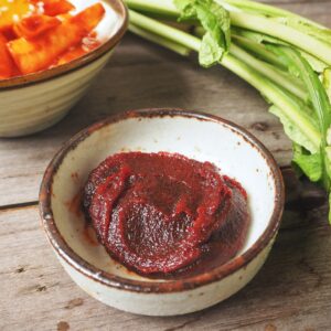 A bowl of gochujang, a dark red paste, next to a bowl of kimchi and some green vegetables.