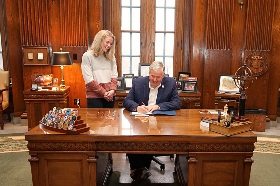 A photo of Grace at standing at the governor's desk, smiling as the Governor, sitting next to her, signs the papers.