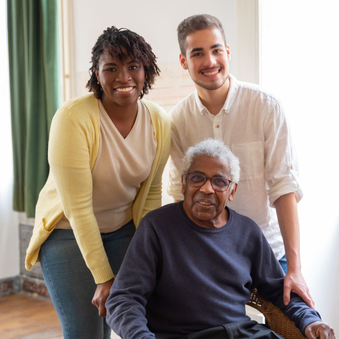 A black woman and man standing behind a sitting elderly black man, all smiling.